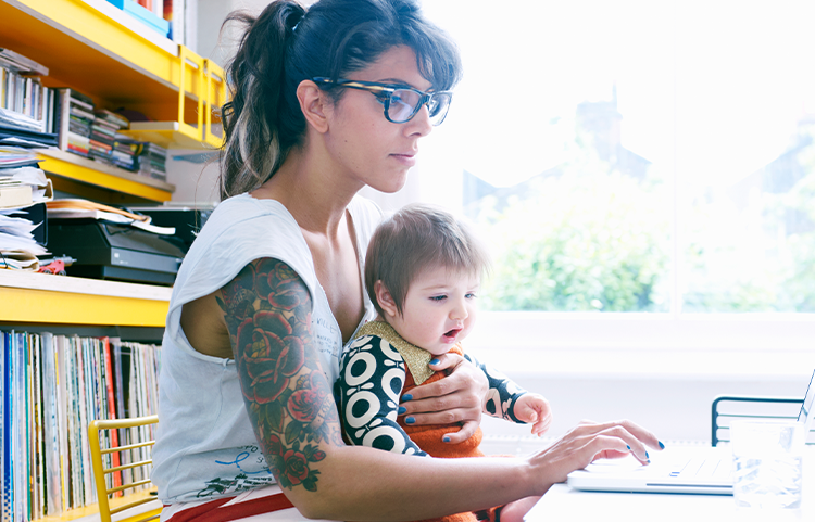 A mother with glasses sits with her baby on her lap while she looks at a laptop
